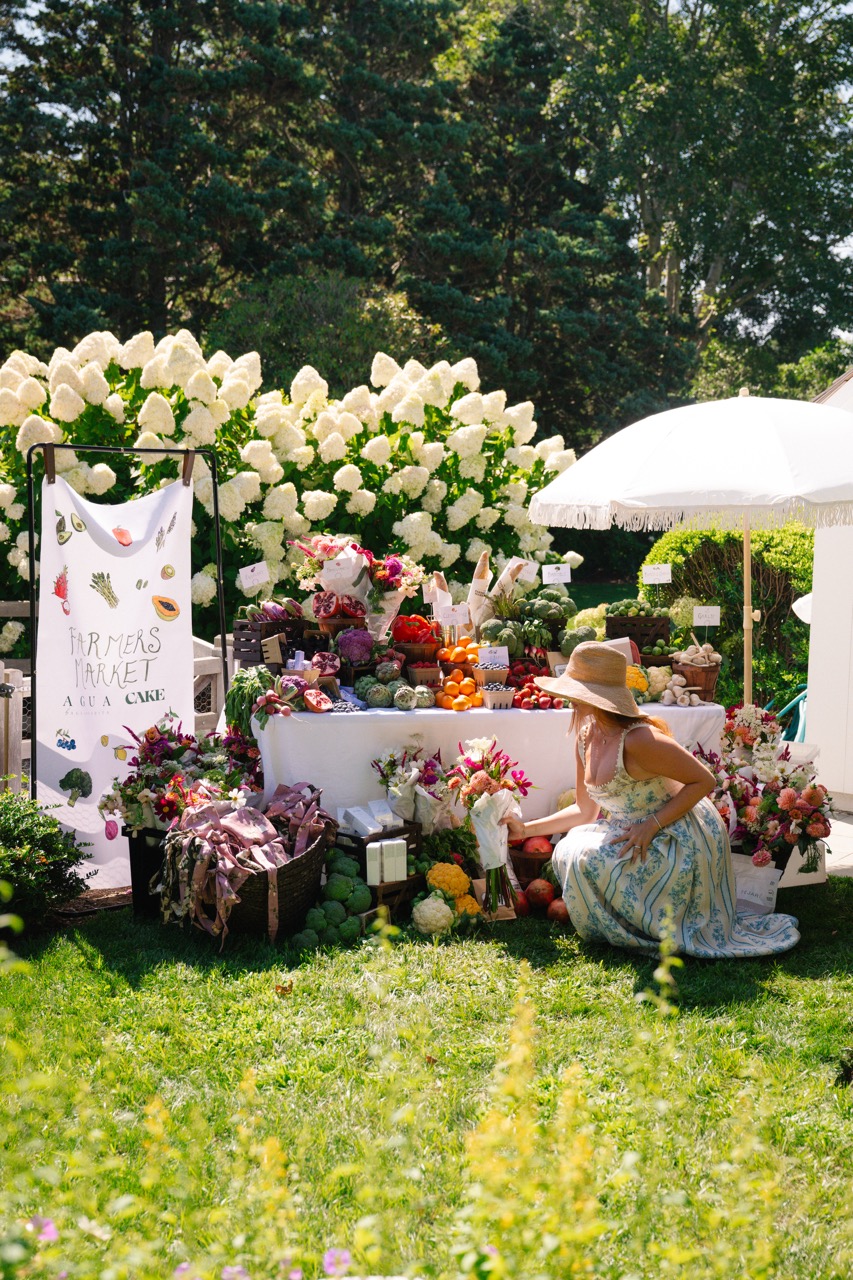 Woman in a floral dress arranging produce and flowers at the Agua by Agua Bendita x CAKE farmer's market brand activation in the Hamptons, featuring a curated outdoor display of fresh produce, florals, and event styling.